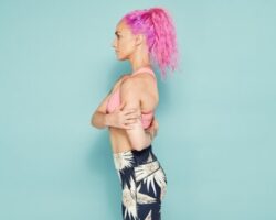 Lady with bright pink hair wearing brightly coloured active wear stand facing away from the camera doing an elbow out rotator stretch