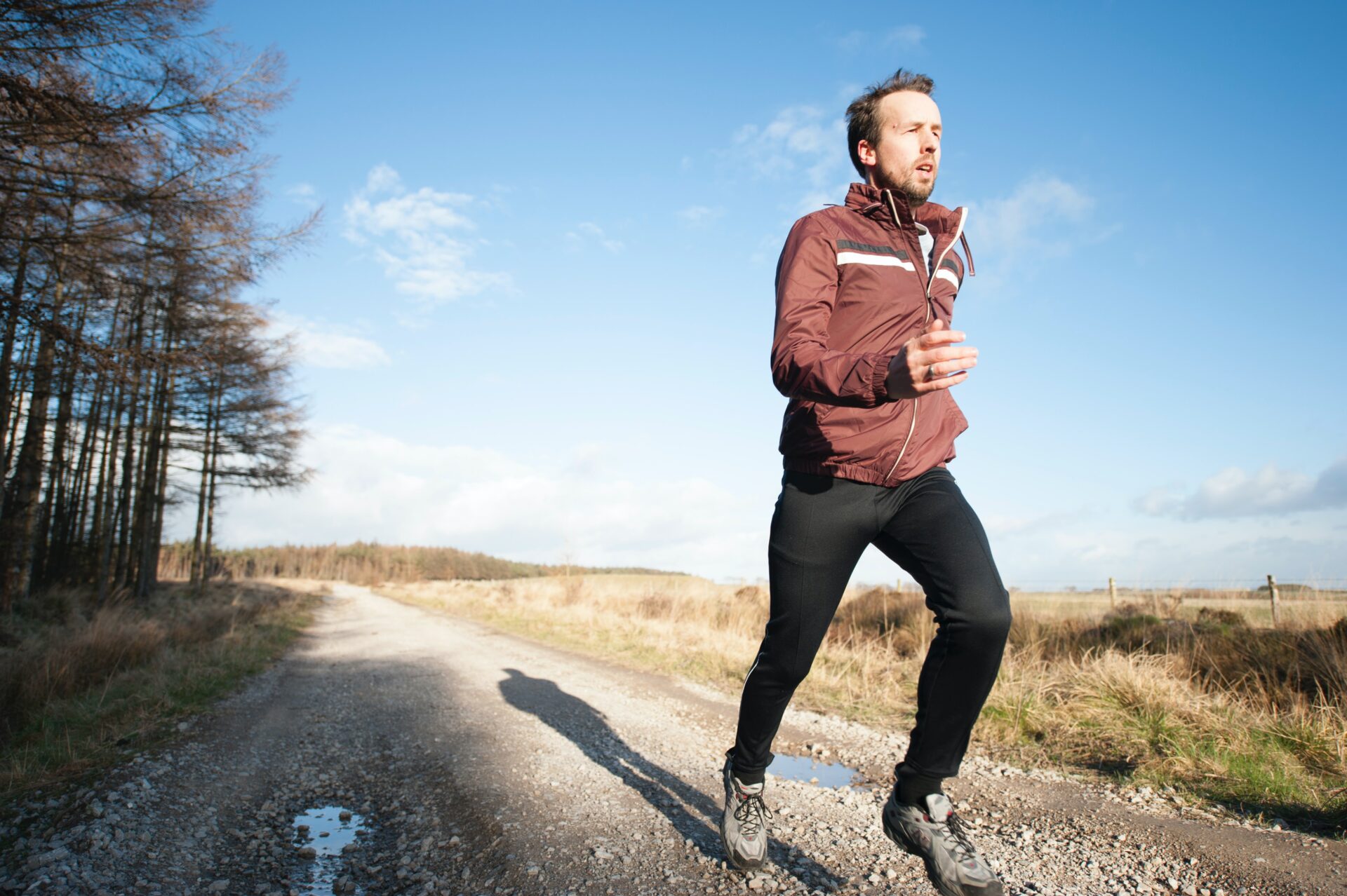 A guy in a brown puffer jacket running on a gravel road in Winter
